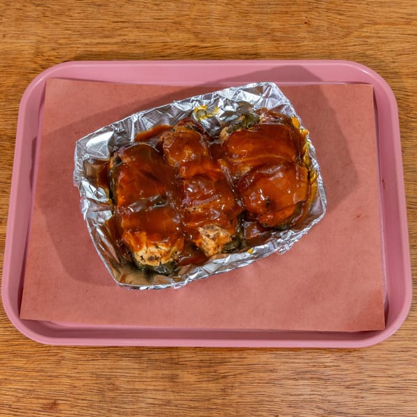 Three saucy BBQ chicken thighs in foil on a pink tray with brown paper, on a wooden table.