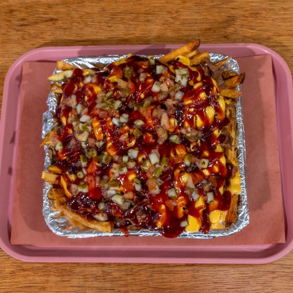 BBQ loaded fries with cheese, BBQ sauce, and diced toppings in a foil container on a pink tray.