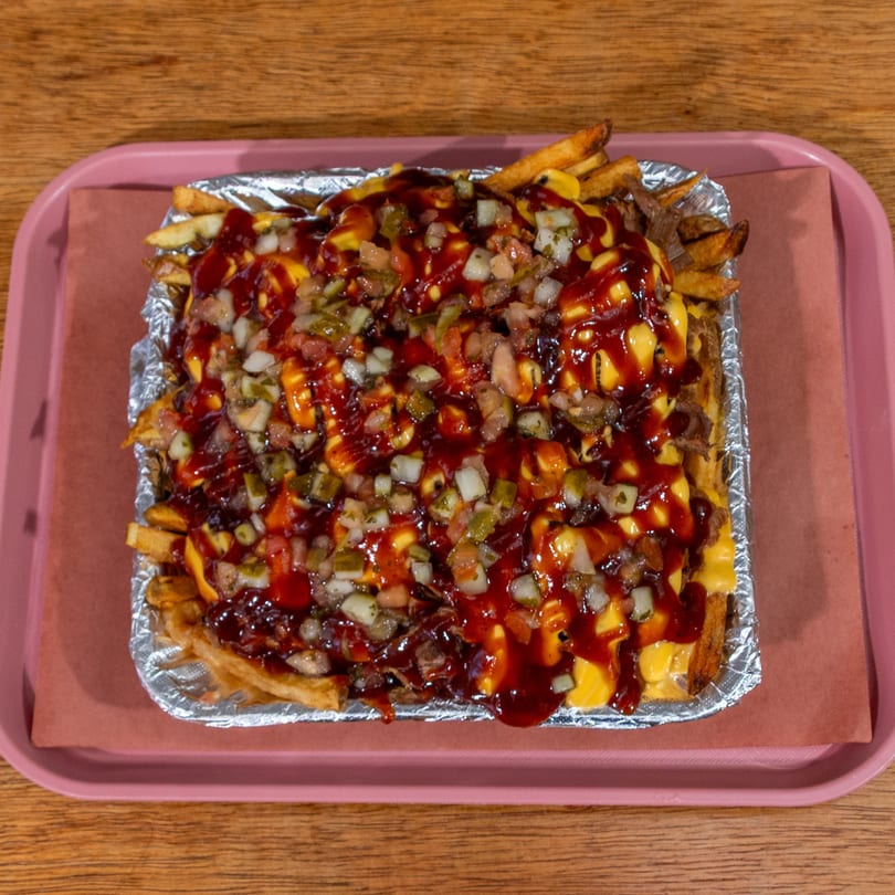 BBQ loaded fries with cheese, BBQ sauce, and diced toppings in a foil container on a pink tray.