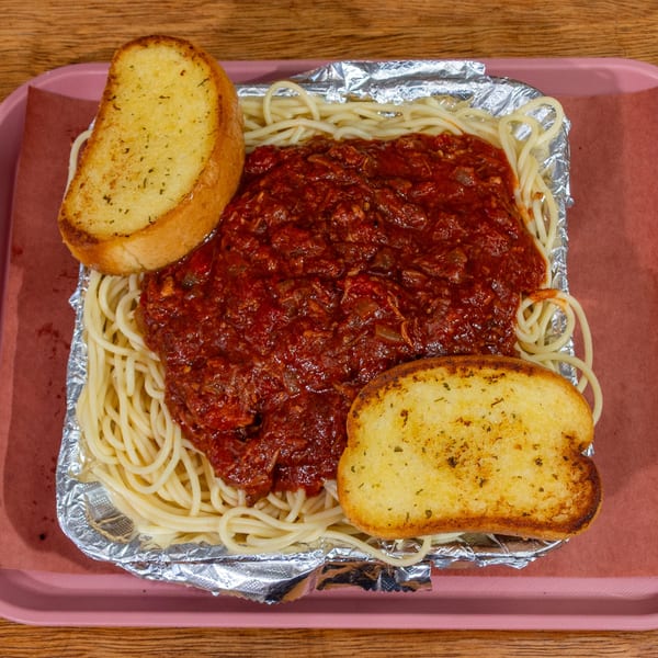Spaghetti with red sauce and two slices of garlic bread in a foil container on a pink tray.